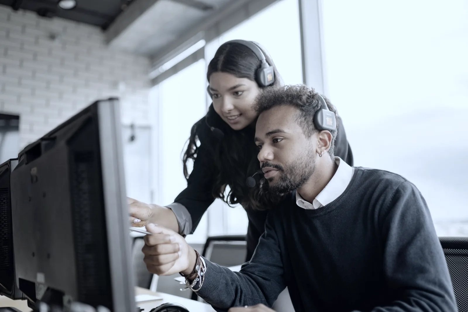 Two service desk technicians wearing headsets look at a screen in a bright open-plan office